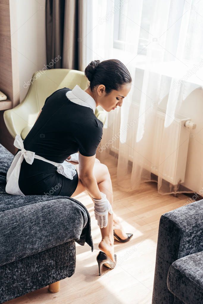 tired maid in white gloves touching legs while sitting on bed