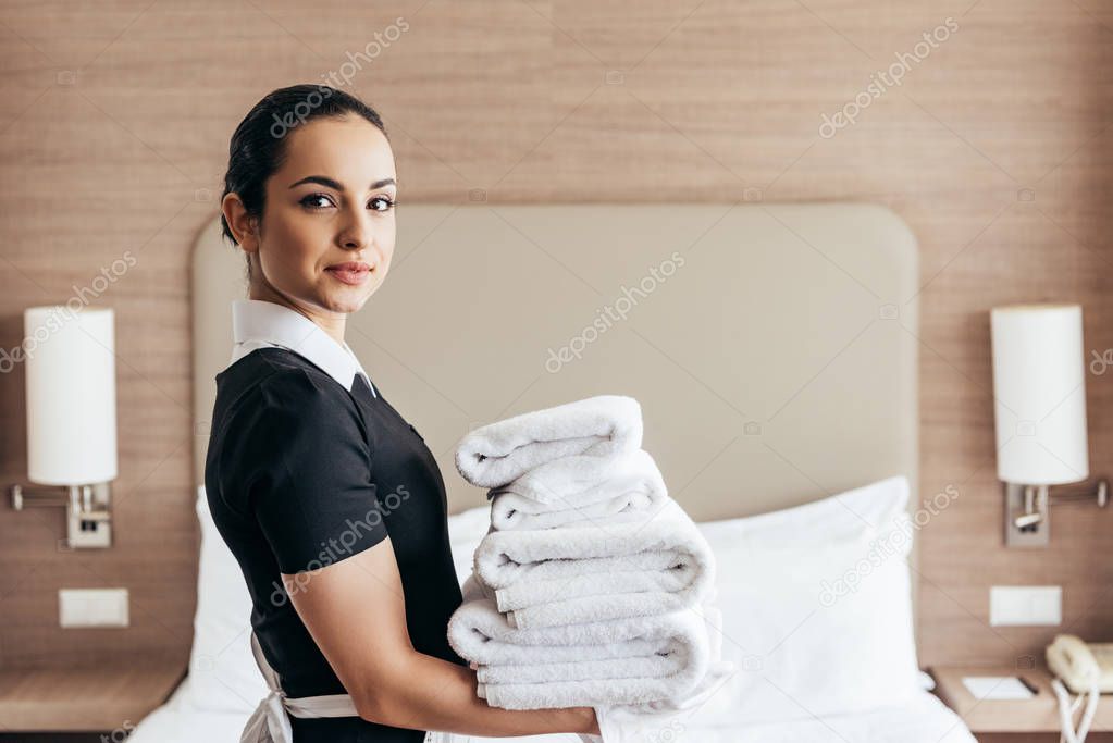 smiling maid holding pile of folded towels near bed and looking at camera in hotel room