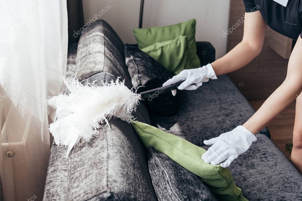 partial view of maid in white gloves cleaning sofa with duster in hotel room