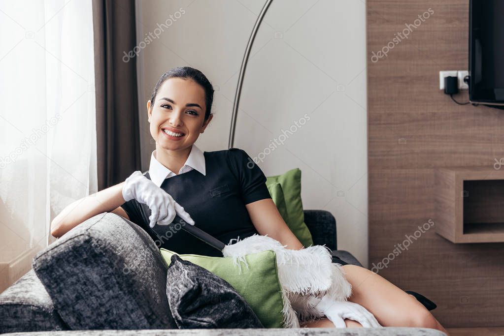 smiling maid in white gloves sitting on sofa and holding duster in hotel room