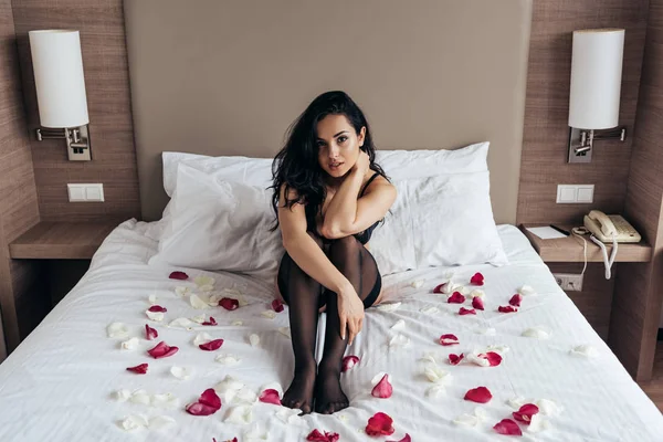 寝室でバラの花びらとベッドに座って黒いストッキングのセクシーなブルネットの女の子 — ストック写真