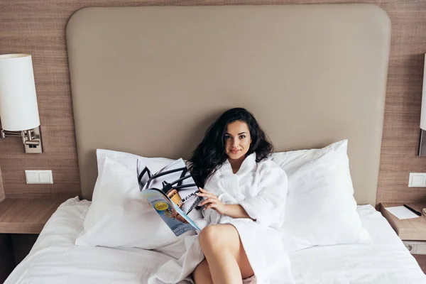 漂亮的黑发年轻女子在白色的浴袍躺在床上和阅读杂志 — 图库照片