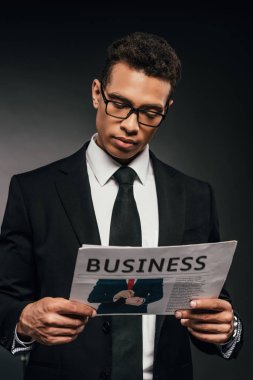 handsome african american businessman in glasses and suit reading business newspaper on dark background clipart
