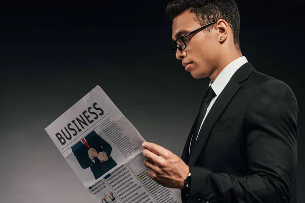 メガネとスーツのハンサムなアフリカ系アメリカ人ビジネスマンのサイドビューは 暗い背景にビジネス新聞を読みます — ストック写真