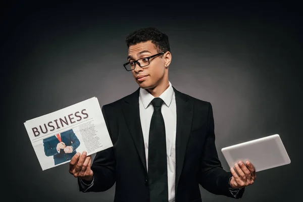 暗い背景にビジネス新聞やデジタルタブレットを保持するメガネとスーツで混乱したアフリカ系アメリカ人のビジネスマン — ストック写真