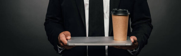 cropped view of african american businessman holding laptop and paper cup on dark background, panoramic shot