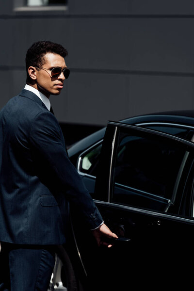 african american businessman in suit and sunglasses opening car door at sunny day