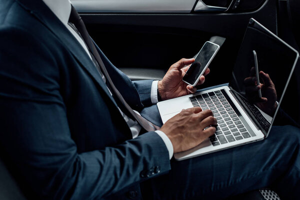 cropped view of african american businessman using laptop and smartphone in car