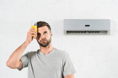 man holding orange juice near forehead and suffering from heat under air conditioner at home clipart