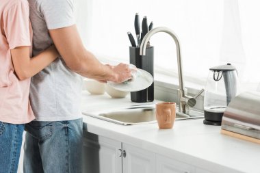 Mutfakta bulaşık yıkayan adama sarılan kadının kırpılmış görünümü