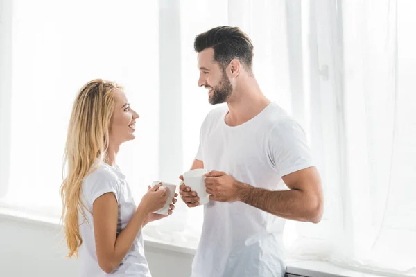 自宅でお互いを見ているコーヒーカップを持つ美しいカップル — ストック写真