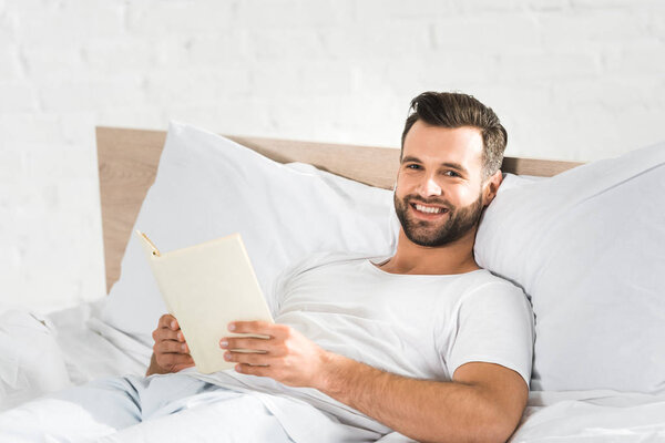 handsome happy man lying in bed and reading book in morning