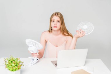 beautiful girl holding electric fan at desk and suffering from heat on grey clipart
