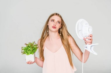 beautiful young woman with Electric Fan and flowerpot suffering from heat isolated on grey clipart
