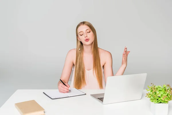 美丽的女孩坐在电脑桌前 在笔记本上写字 在灰色上遭受热 — 图库照片