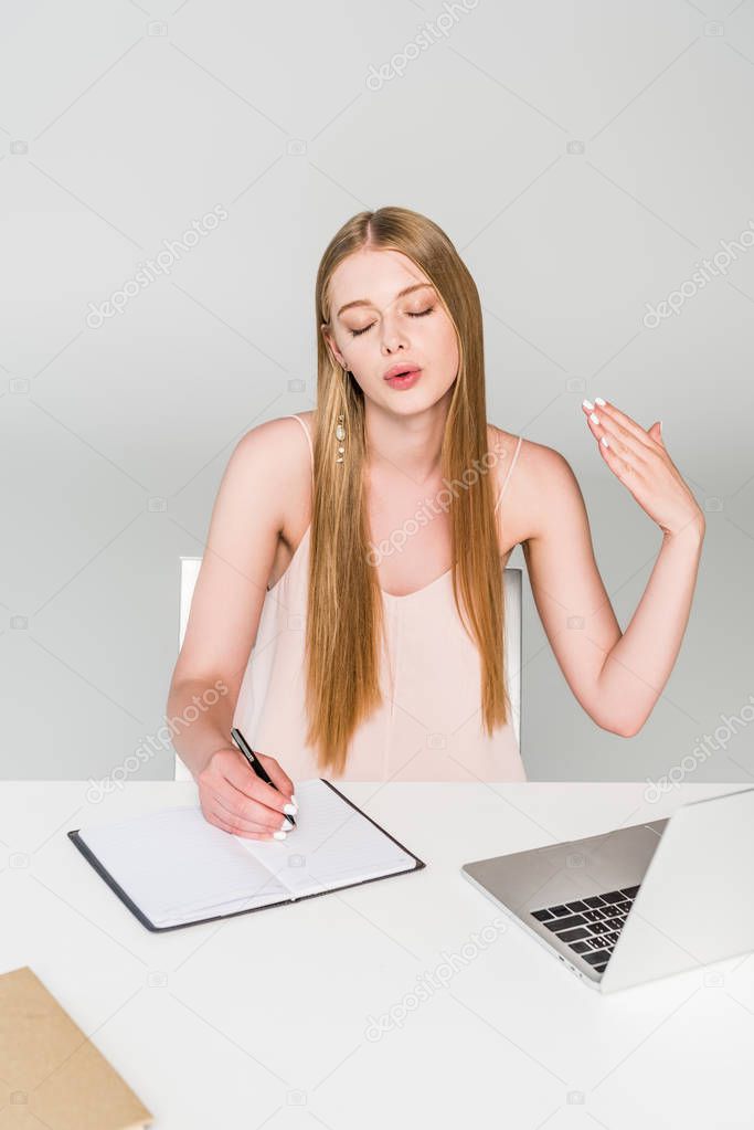 beautiful girl sitting at computer desk, writing in notebook and suffering from heat on grey