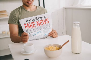 kahve ve kase gevreği ile fincan yakın mutfak masasında otururken Asyalı adam sahte haber gazetesi okuma kırpılmış görünümü