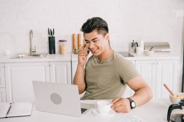 Laptop yakın mutfak masasında otururken akıllı telefonda konuşan neşeli Asya adam