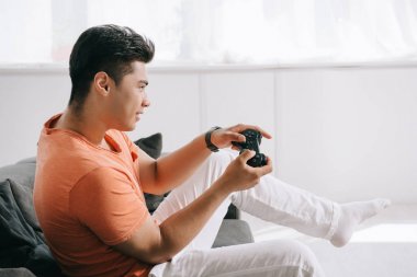 Kiev, Ukrayna-Haziran 12, 2019: Genç Asyalı adam joystick ile video oyunu oynarken evde kanepede otururken.