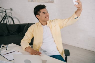 evde masada otururken akıllı telefon ile selfie alarak gülümseyen Asya adam