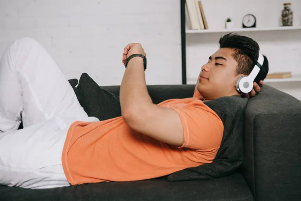 young asian man looking at watch while lying on sofa and listening music in headphones