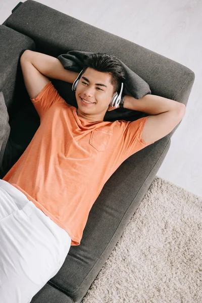 üst görünüm gülümseyen Asya adam kanepede yatarken ve kulaklık müzik dinleme