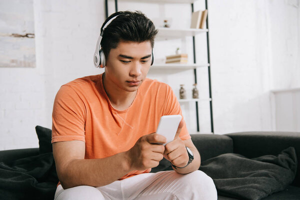 thoughtful asian man listening music in headphones and using smartphone while sitting on sofa at home