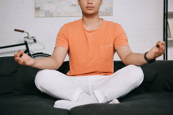обрезанный вид молодого человека медитирующего сидя на диване в позе лотоса
