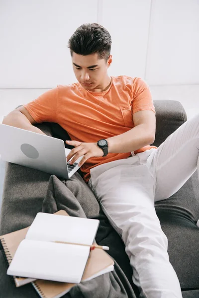 ノートの近くのソファに座っている間 ラップトップを使用する若いアジアの男性 — ストック写真