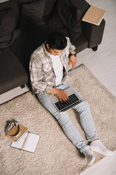 overhead view of young man sitting on floor near couch and using laptop at home
