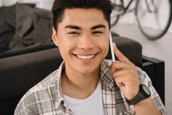 Young Cheerful Asian Man Smiling Camera While Talking Smartphone Home — Stock Photo, Image