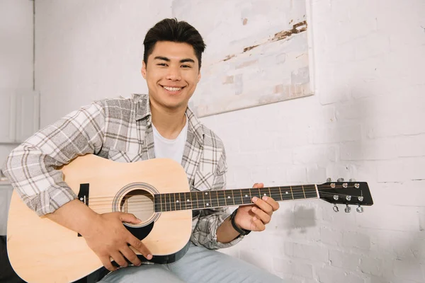 Feliz Asiático Hombre Sonriendo Cámara Mientras Jugando Acústica Guitarra Casa — Foto de Stock