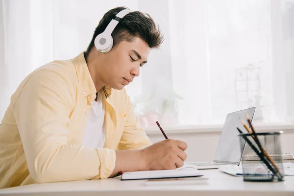 Jovem Concentrado Asiático Homem Fones Ouvido Escrevendo Notebook Branco Sentado — Fotografia de Stock