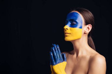 naked young woman with painted Ukrainian flag on skin praying with closed eyes isolated on black clipart
