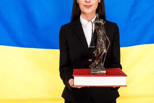 cropped view of lawyer in black suit holding Themis figurine on book on Ukrainian flag background