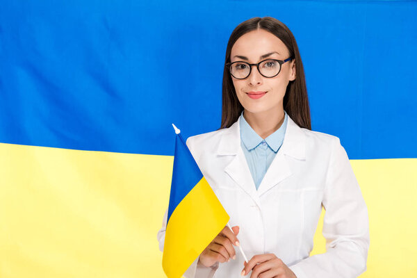 smiling doctor in white coat and glasses on Ukrainian flag background
