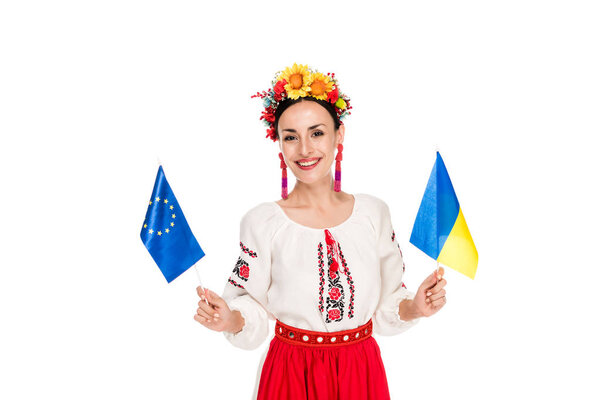 happy brunette young woman in national Ukrainian costume holding European and Ukrainian flags isolated on white