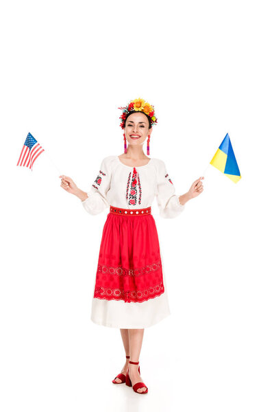 happy brunette young woman in national Ukrainian costume holding American and Ukrainian flags isolated on white