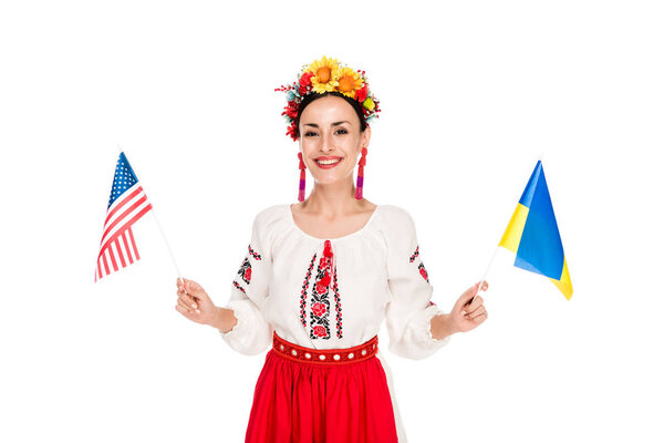 smiling brunette young woman in national Ukrainian costume holding American and Ukrainian flags isolated on white