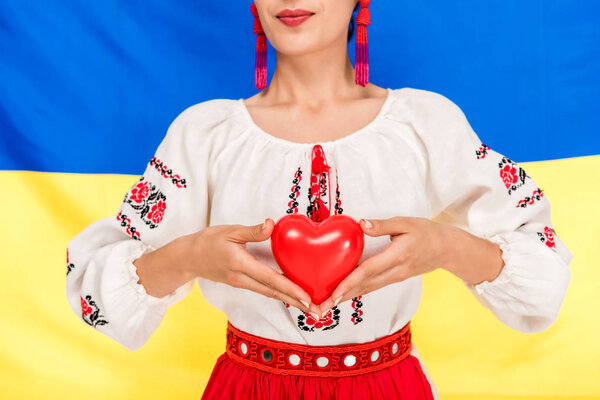 partial view of young woman in national Ukrainian costume holding red heart with flag of Ukraine on background