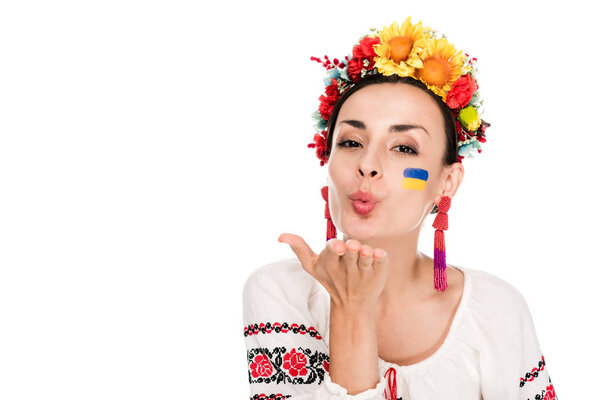 brunette young woman in national Ukrainian embroidered shirt and floral wreath blowing kiss isolated on white
