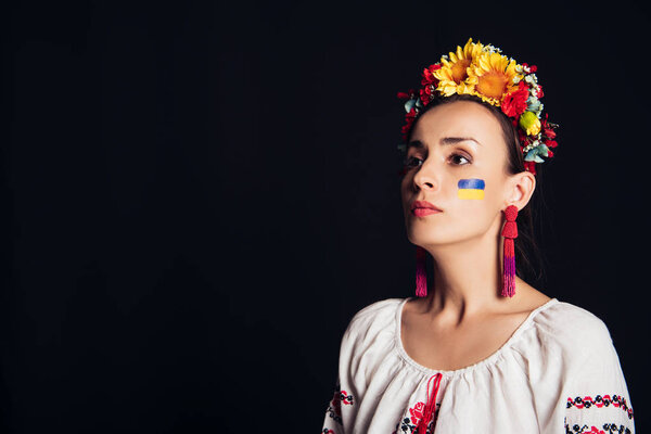 brunette young woman in national Ukrainian costume and floral wreath looking away isolated on black