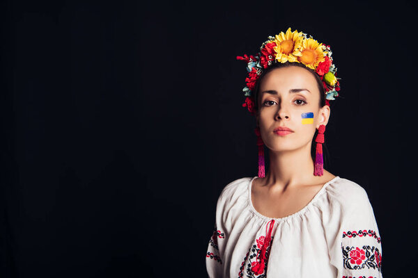 brunette young woman in national Ukrainian costume and floral wreath looking at camera isolated on black