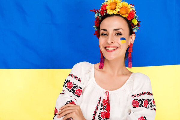 happy brunette young woman in national Ukrainian costume and floral wreath with flag of Ukraine on background