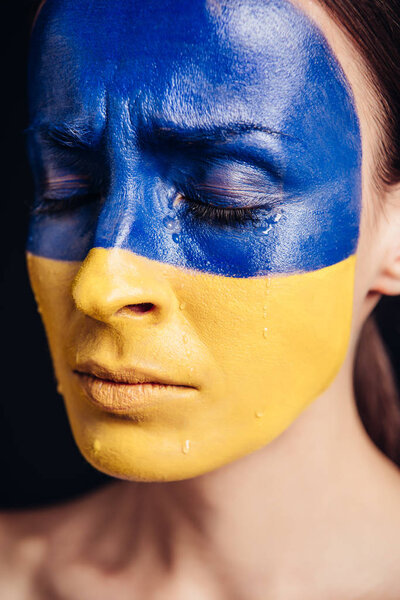 close up view of young woman with painted Ukrainian flag on skin crying isolated on black