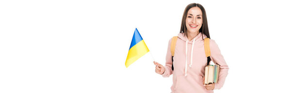 smiling student with backpack holding Ukrainian flag and books isolated on white, panoramic shot