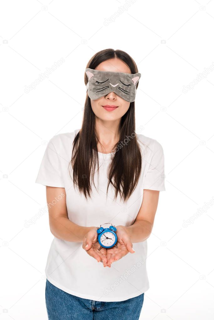 brunette young woman in cat sleeping eye mask holding toy alarm clock isolated on white