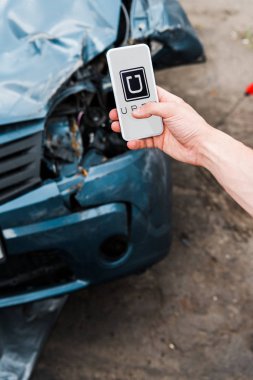 Kiev, Ukrayna - 20 Haziran 2019: hasarlı arabanın yanında ekranda uber uygulaması ile akıllı telefon tutan adamın kırpılmış görünümü 