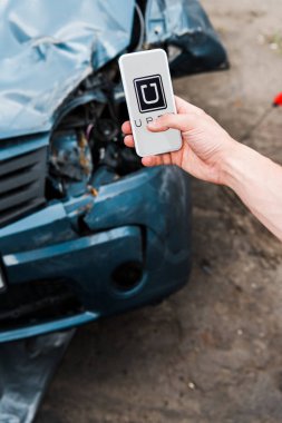 KYIV, UKRAINE - JUNE 20, 2019: cropped view of man holding smartphone with uber app on screen near damaged car  clipart