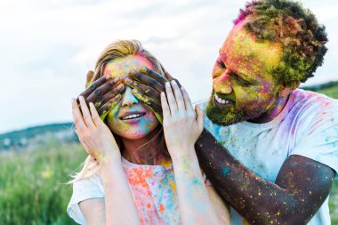 mutlu afrikalı amerikalı adam yüzünde holi boyalar ile genç kadının gözlerini kaplayan 